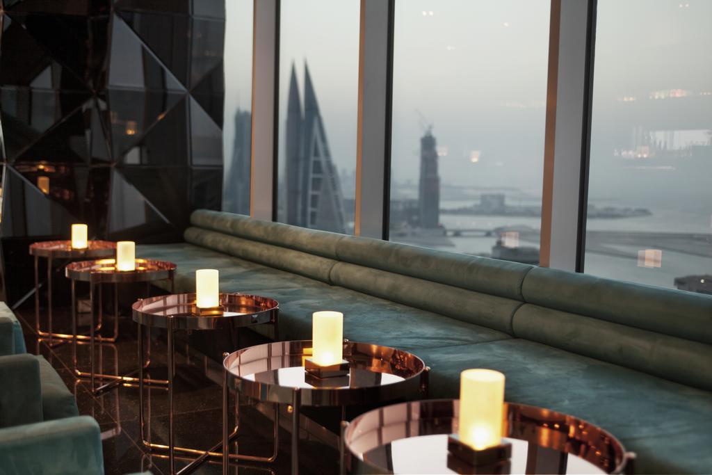 The Domain Bahrain Hotel And Spa - Adults Friendly 16 Years Plus 麦纳麦 外观 照片 The photo depicts a stylish lounge area with a modern aesthetic. There are several circular tables made of metallic materials, each topped with a small glowing candle. A long, plush seating area in a teal color runs along the window, offering a view 